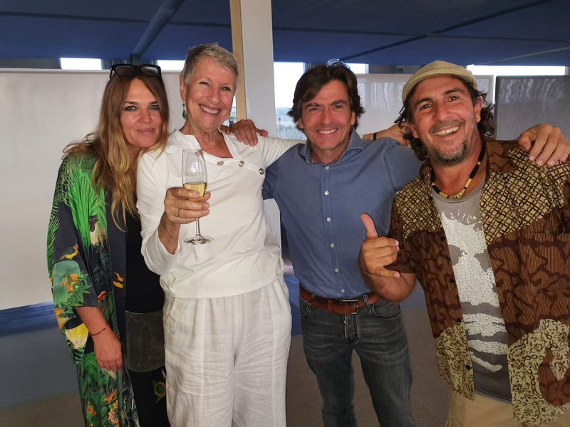 Four people in a row, smiling with arms around each other.Two women on the left, second to left is holding a glass of champagne or white wine. The man on the far right has a cap on and his making the "shaka" sign with his right hand. 